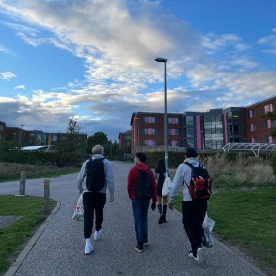 A group of students wearing backbacks and carrying bags walk towards a university building with a cloudy blue sky in the background