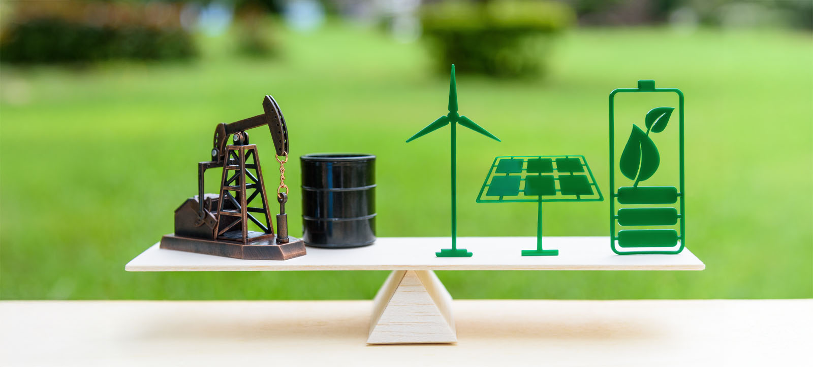 Different energy sources - fossil fuels, wind power, solar power, and electric - are lined up on a piece of wood, which is precariously balanced on a pivot. The background is a green field in soft focus.