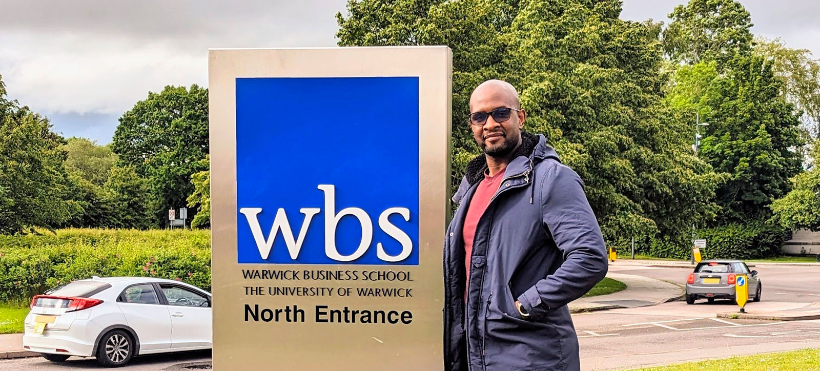 Robert Ndebele stands next to the Warwick Business School logo outside the School Robert Ndebele stands next to the Warwick Business School logo outside the School