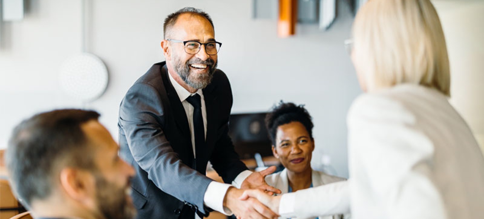 A B2B salesperson shakes hands with a customer after agreeing a sale. They are each accompanied by a member of their respective teams in an office setting. A B2B salesperson shakes hands with a customer after agreeing a sale. They are each accompanied by a member of their respective teams in an office setting.