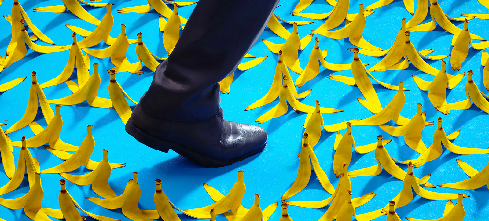 A close up of the foot and lower leg of a businessperson in a suit as they walk through a minefield of banana skins of the floor around them.