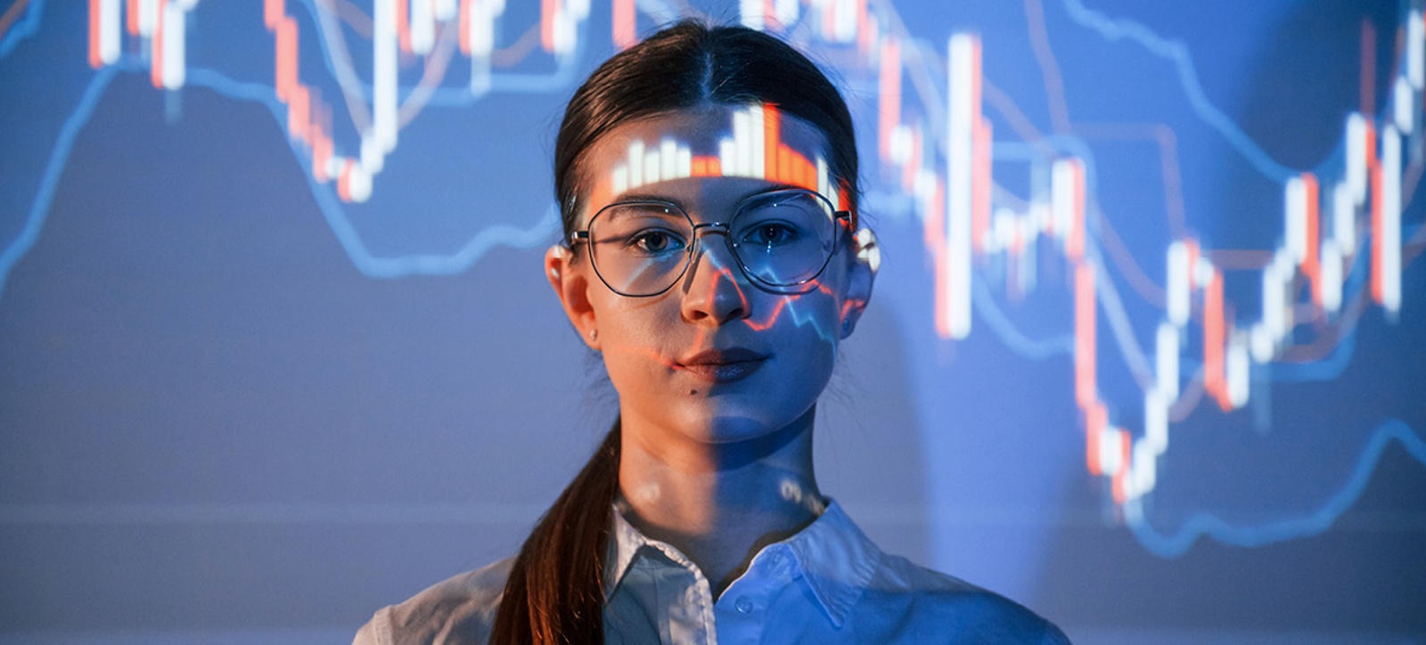 A young business leader looks to the camera. Financial data is projected onto her face and a screen behind her.