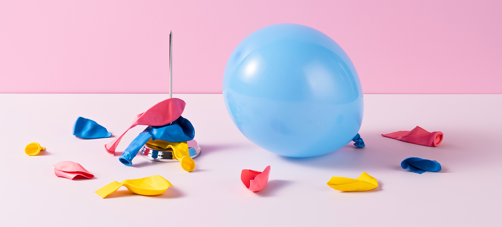 A light blue balloon sits on a table top next to a tall metal spike filled with the remnants of brightlycoloured burst balloons. A light blue balloon sits on a table top next to a tall metal spike filled with the remnants of brightlycoloured burst balloons.