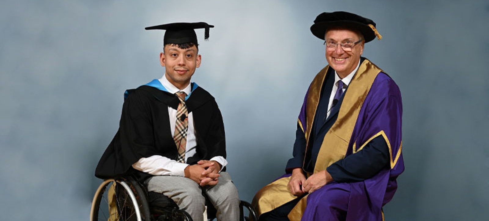 Warwick Business School Sami Dar, wearing a graduation cap and gown, is sat in his wheelchair next to University of Warwick Vice Chancellor Stuart Croft, who is also wearing ceremonial robes and sitting down.
