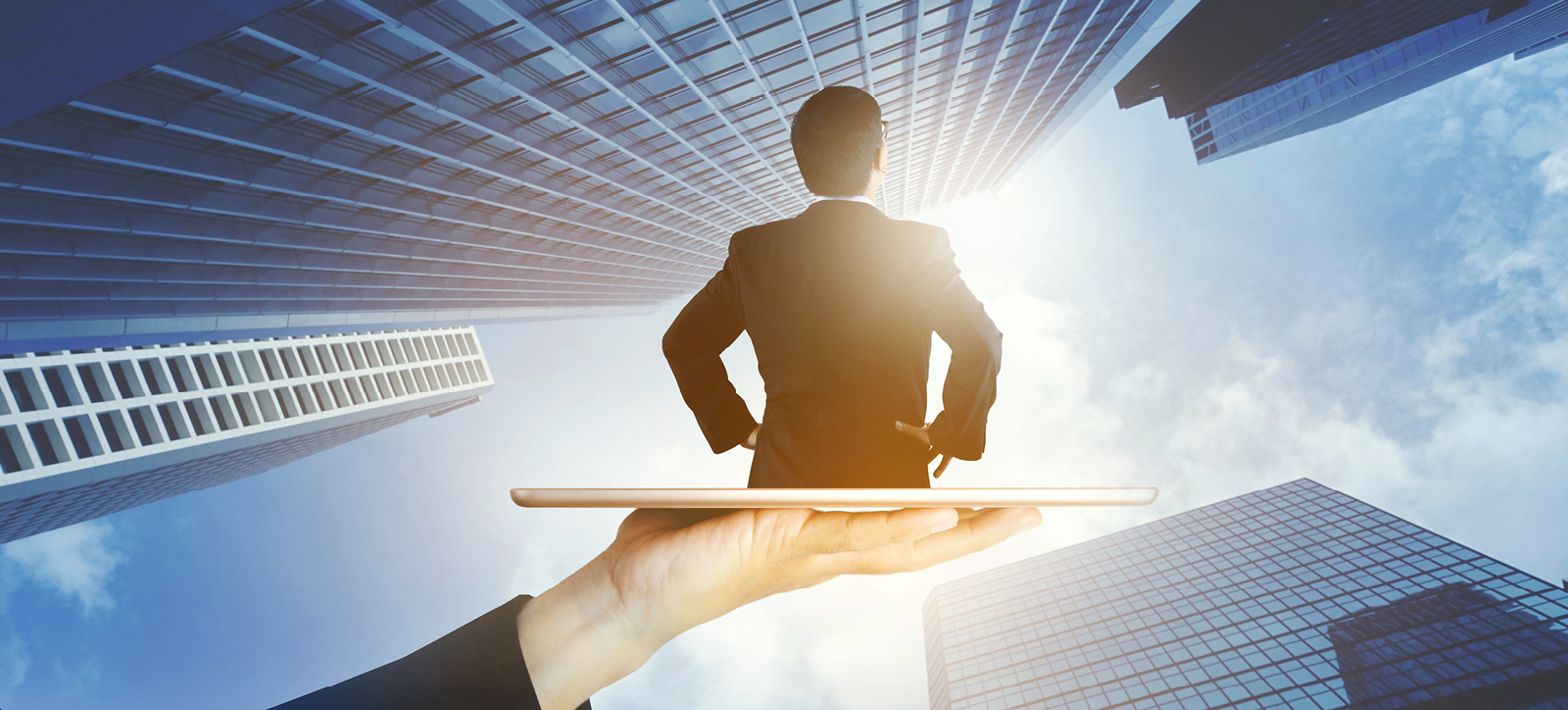 The upper half of a businessman is seen emerging from a tablet screen. The tablet is being held up by a giant hand and arm dressed in a suit jacket. The background is a photograph of skyscraper office blocks taken looking upwards towards the sky.