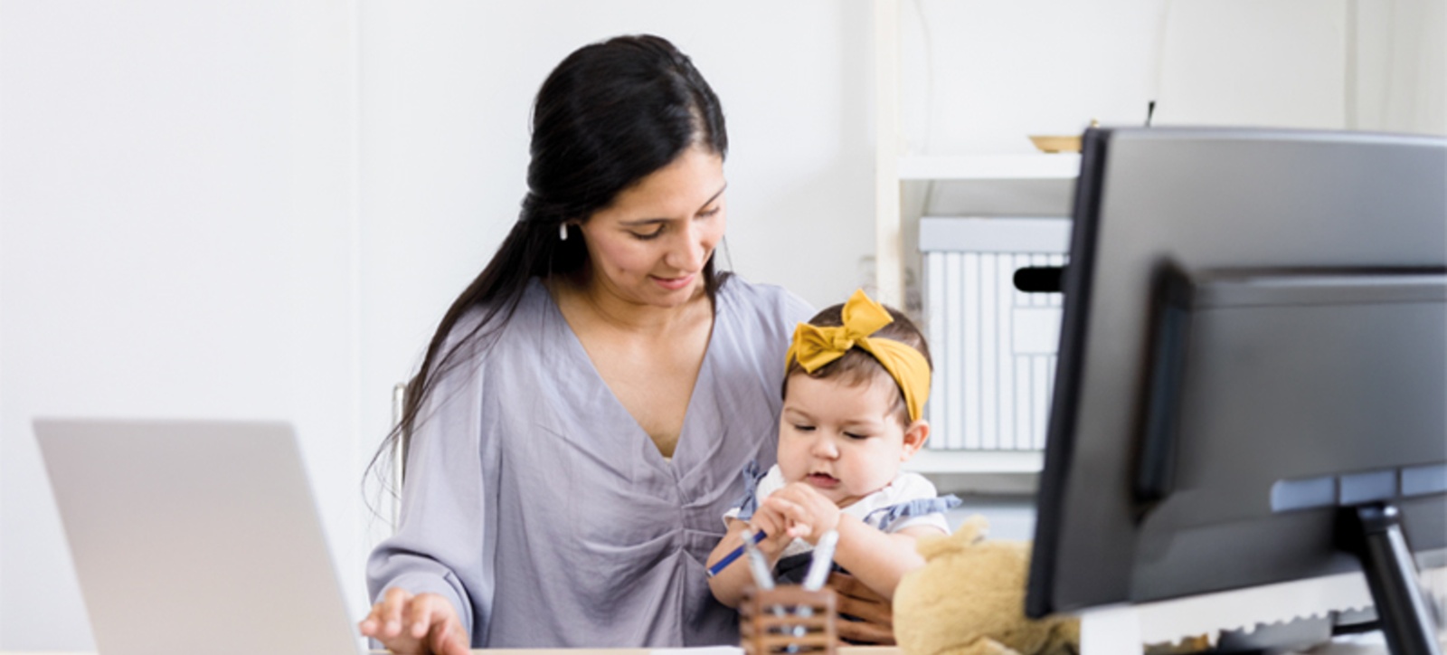 A hybrid worker sits at her computer at home with her baby on her lap while she works. A hybrid worker sits at her computer at home with her baby on her lap while she works.