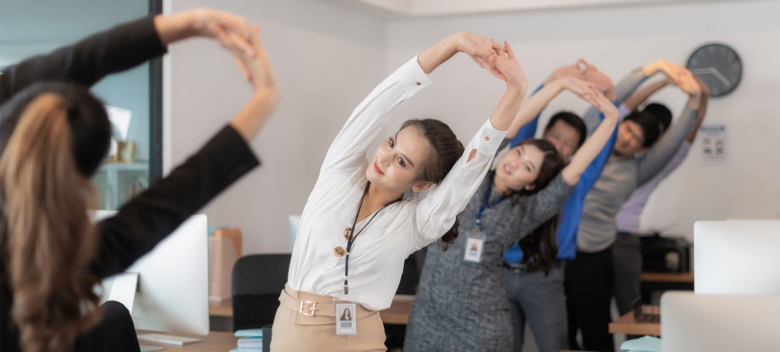 A row of office workers participate in stretching exercises at their desks, following a team leader at the front who is guiding them.