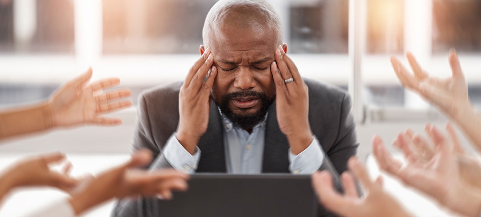 A overwhelmed-looking businessman sits at the head of the meeting table. His is resting his elbows on the desk and has his hands on his temples and he shuts his eyes in frustration. In the foreground a number of employees hold out their hands towards him, asking him to complete tasks or provide answers.