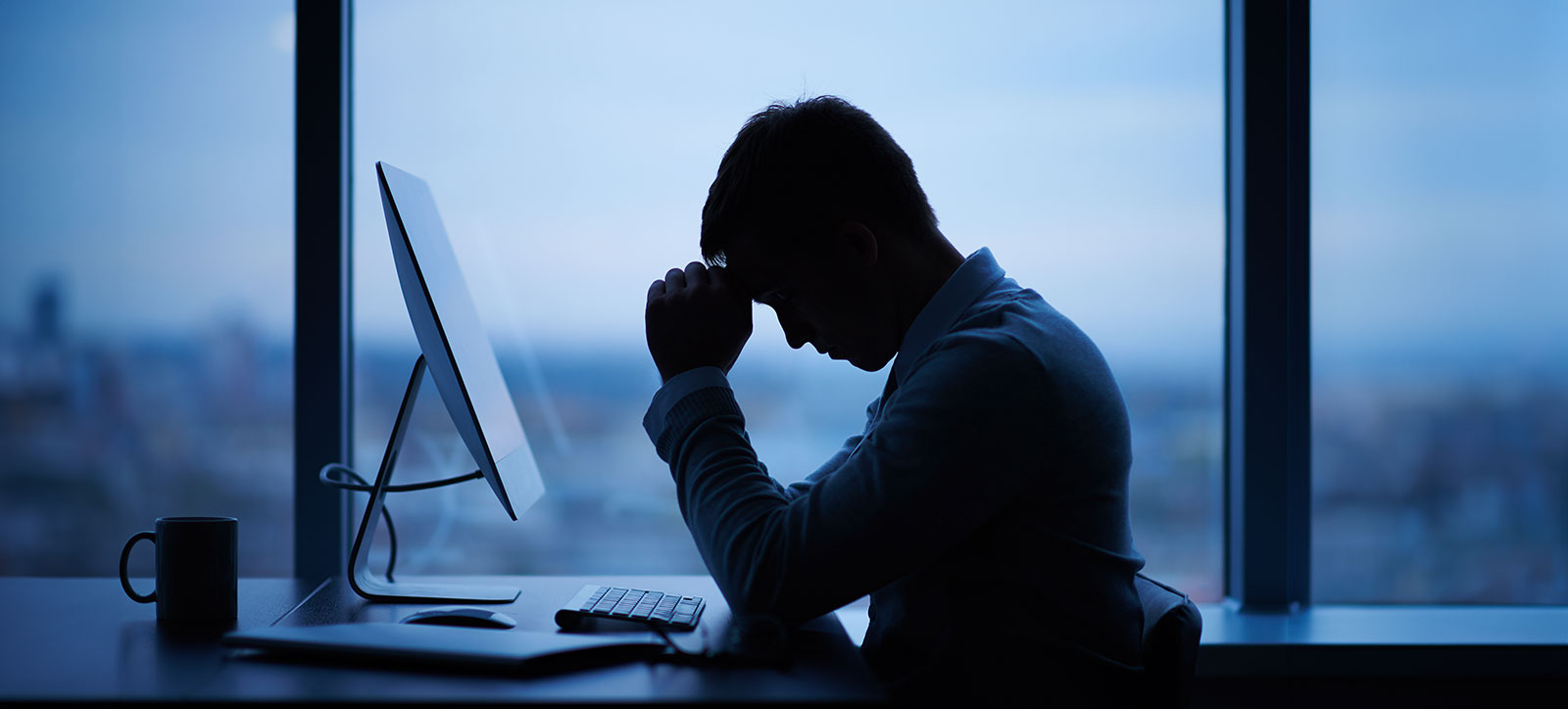 A male office worker in shadow with his head in his hands at his desk A male office worker in shadow with his head in his hands at his desk
