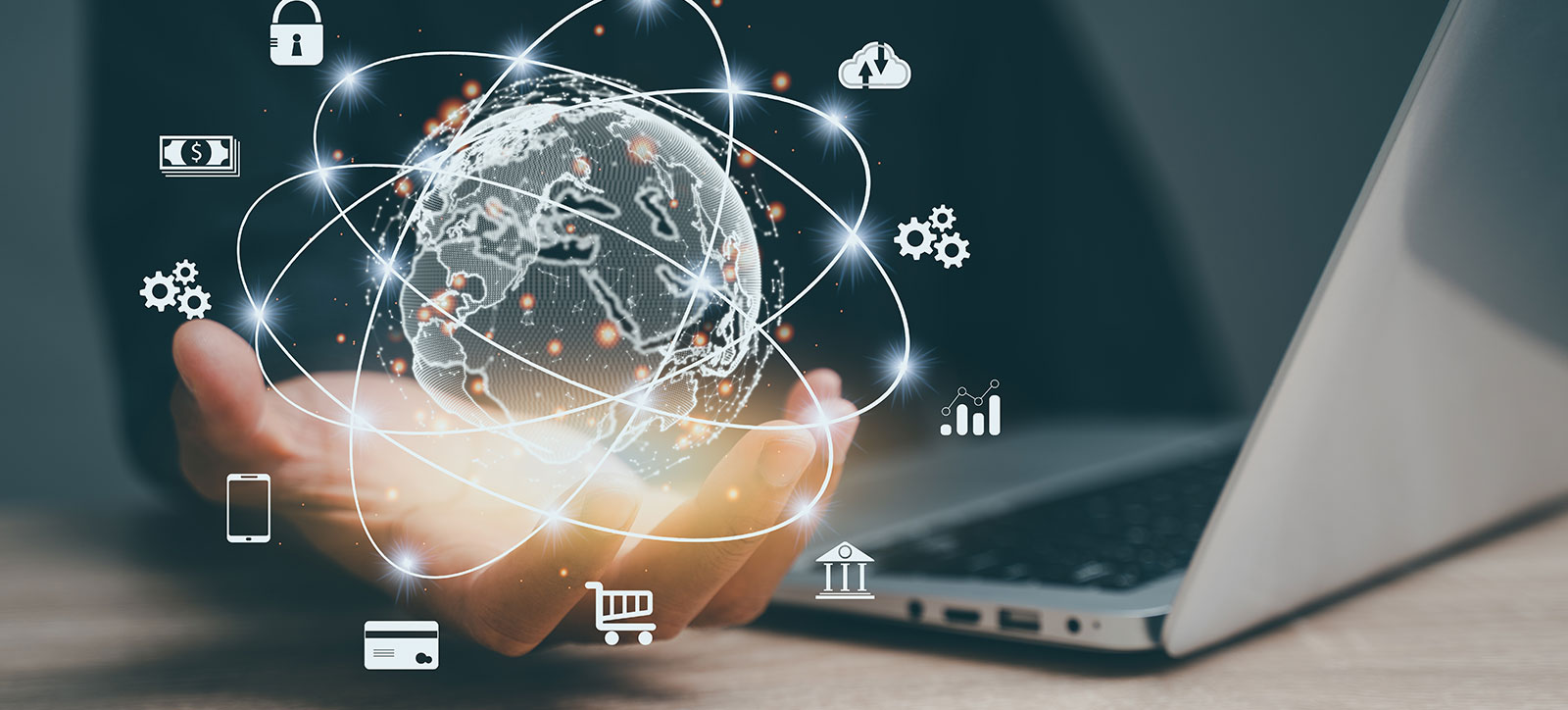 A close up shot of a worker sat in front of a laptop computer on a desk. They are holding out their right hand, which rests on the table, with their open palm facing upwards. Their hand is 'holding' a virtual image of planet Earth, encircled by a series of rings so it looks like an atom. Around the planet are a series of icons such as a padlock, a stack of dollar bills, a credit card, a bank, a mobile phone, and a data cloud to illustrate a fintech ecosystem. A close up shot of a worker sat in front of a laptop computer on a desk. They are holding out their right hand, which rests on the table, with their open palm facing upwards. Their hand is 'holding' a virtual image of planet Earth, encircled by a series of rings so it looks like an atom. Around the planet are a series of icons such as a padlock, a stack of dollar bills, a credit card, a bank, a mobile phone, and a data cloud to illustrate a fintech ecosystem.