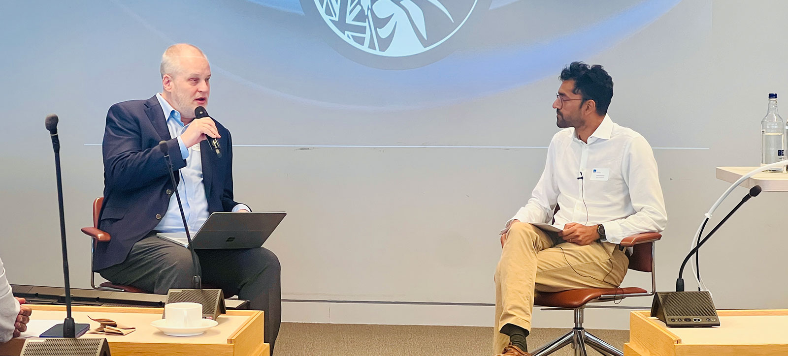 Two men in discussion seated in front of a screen showing the logo of the Bank of England, Britannia, the female personification of the British Isles.