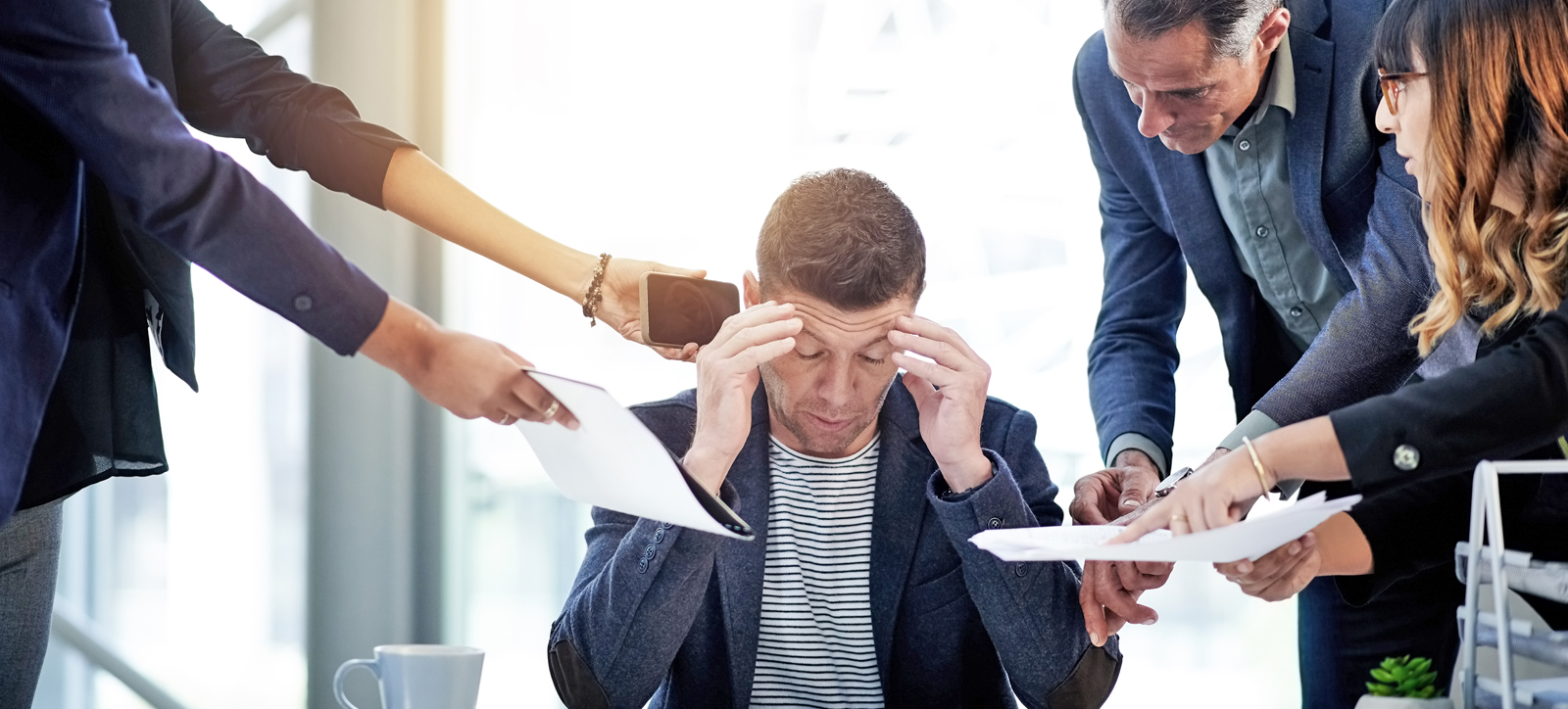 A manager sits with his hands to his temples, struggling with doubt as senior colleagues surround him with questions.