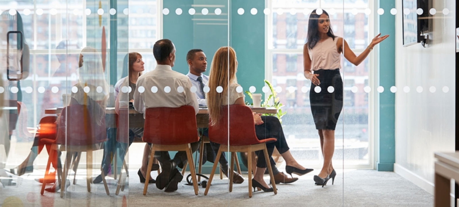 A women stood in an office, dressed smartly and pointing at a screen while delivering a presentation to a group of people sat on a table.