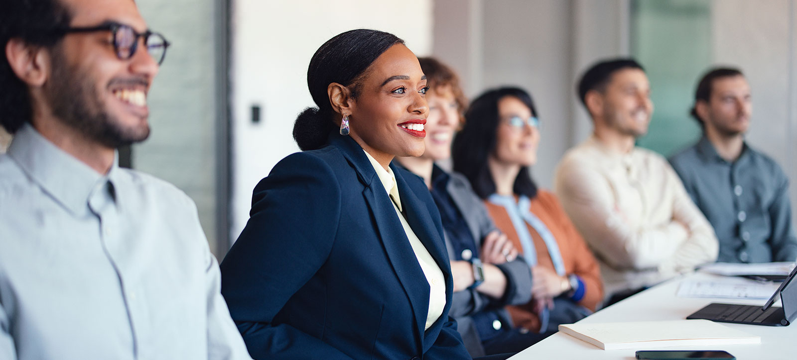 A diverse group of professionals sits in a bright office, listening and smiling as a confident woman in a navy blazer leads the presentation. Laptops and documents on the table suggest a collaborative meeting.