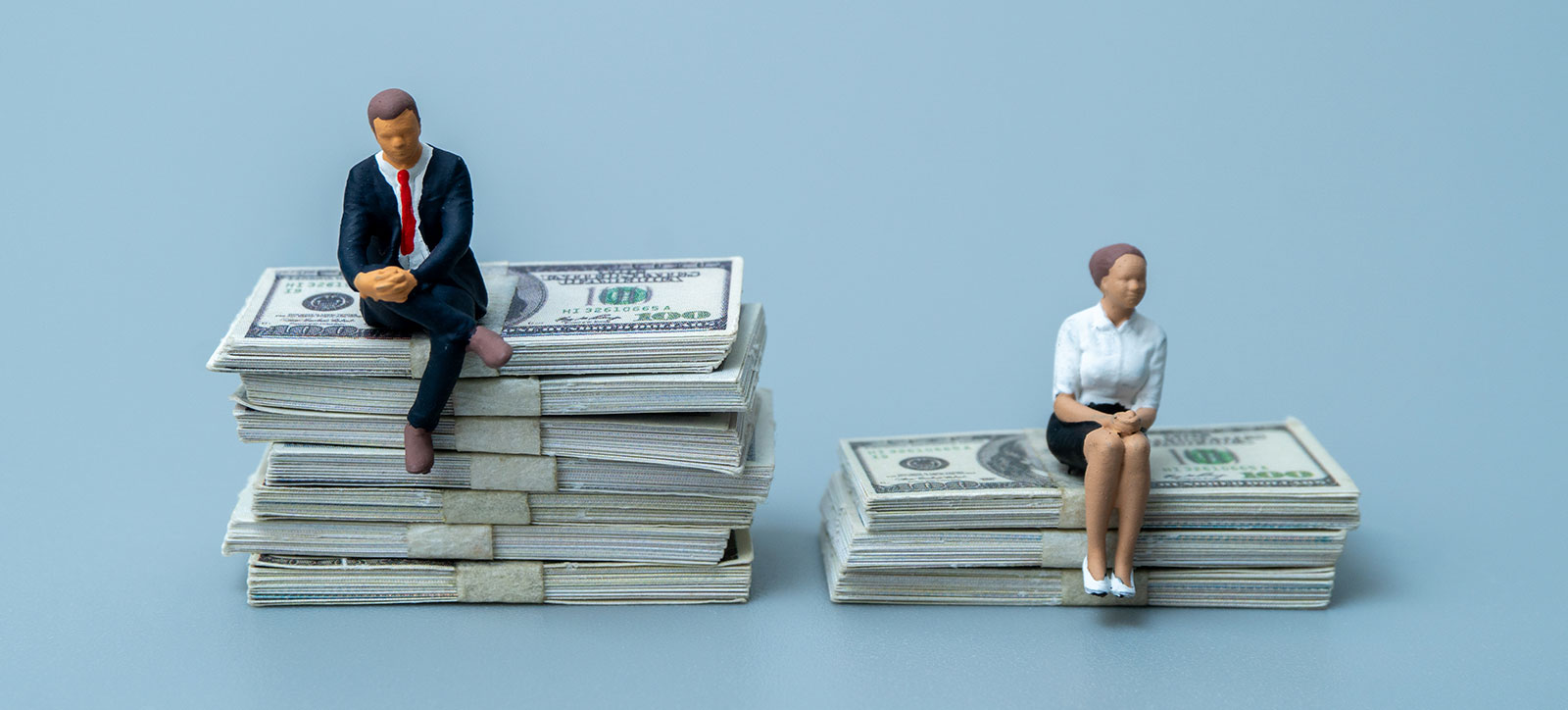 A model of a woman in a business suit sits on a small pile of $100 bills, while a model of a man in a business suit sits on a much bigger pile.