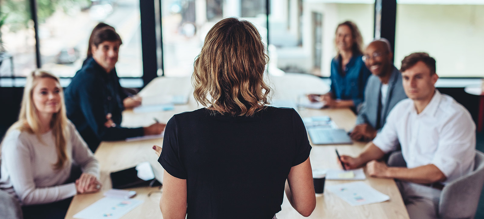 A blonde woman presenting in front of six people around a boardroom table