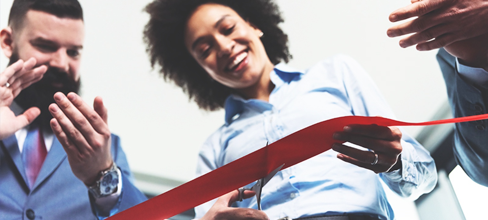 A young female entrepreneur, smartly dressing in a light blue shirt and grey skirt, cuts a bright red ribbon to open a new business. A young businessman in a suit, with short hair and a bear, stands next to her applauding.
