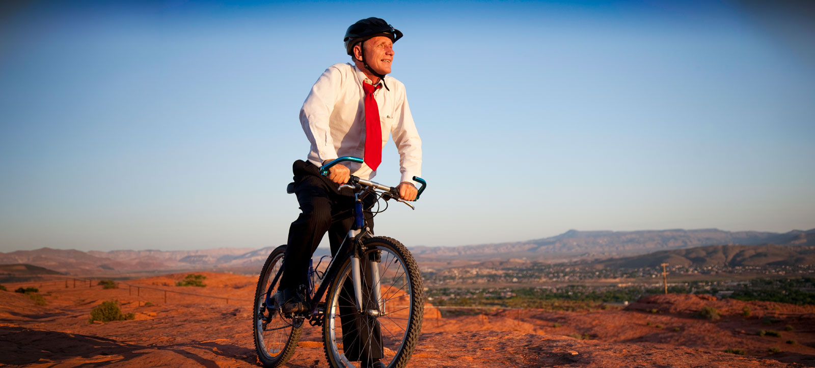 A businessman in a white shirt, red tie, and dark trousers sits on a bicycle at the crest of a hill, looking out across the hot and arid landscape that surrounds him.