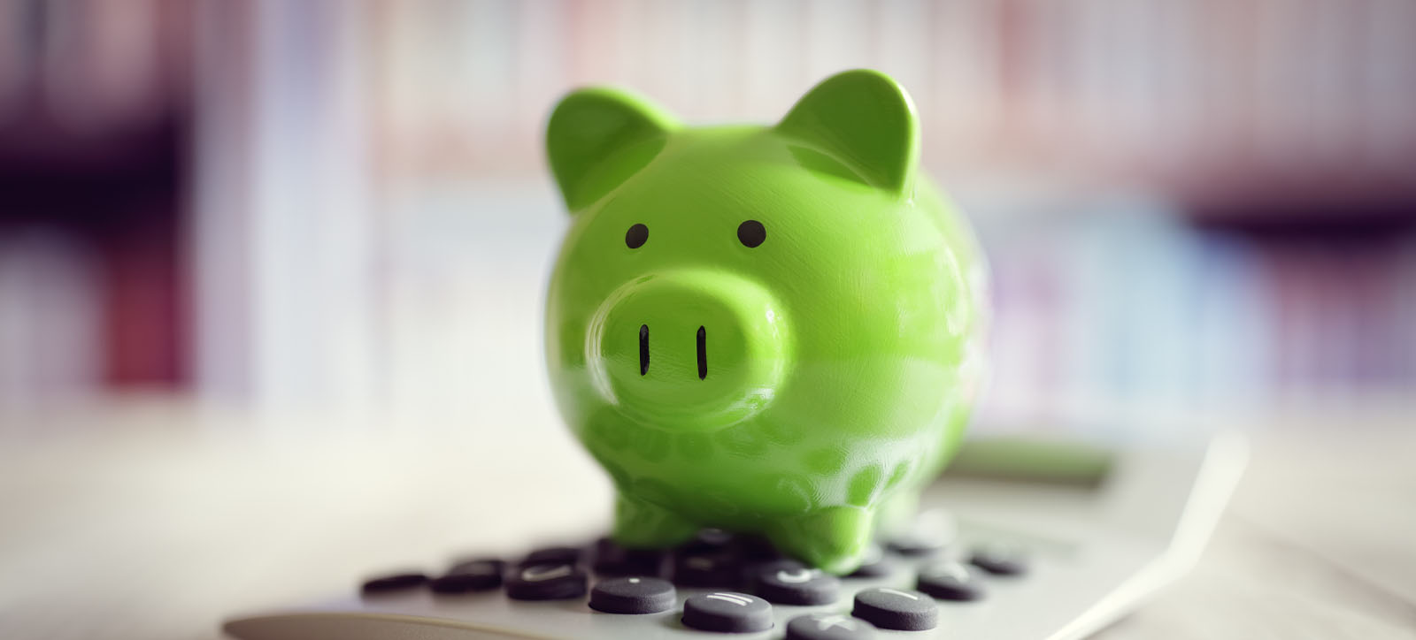 A green piggy bank sits on a calculator on a desk. A bookcase is visible in soft focus in the background