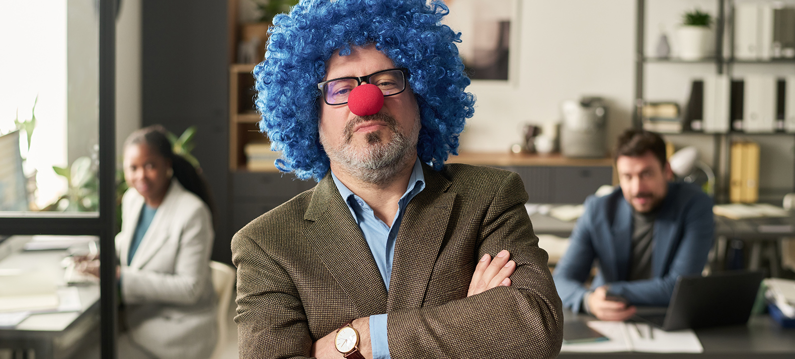 A male CEO in a suit is wearing a blue clown wig and red nose. He has his arms crossed and is frowned, like his joke has failed to amuse the workers in the background of the photograph
