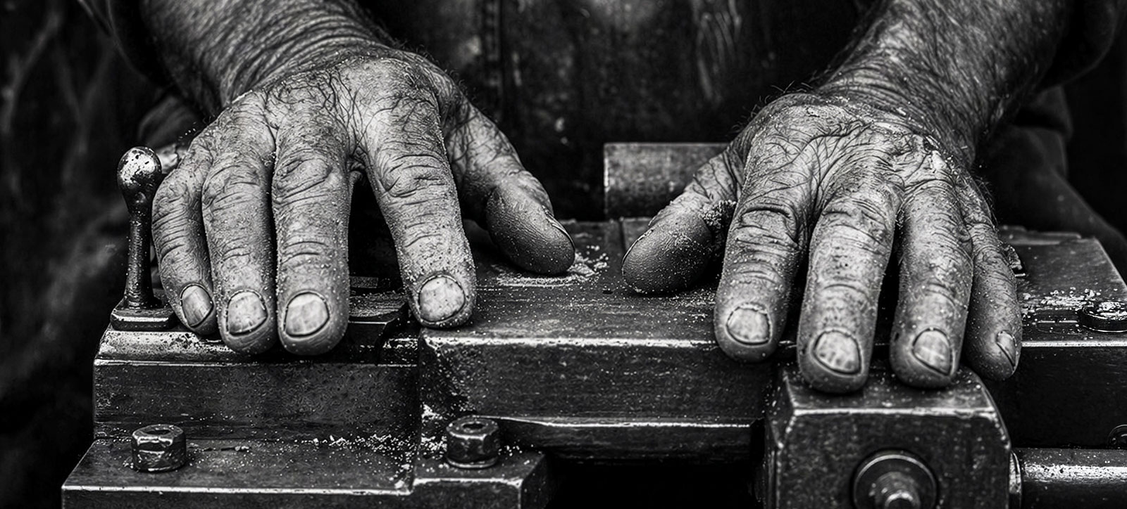 A close up black and white photograph of a pair of workman's weathered, wrinkled hands as they rest on a workbench