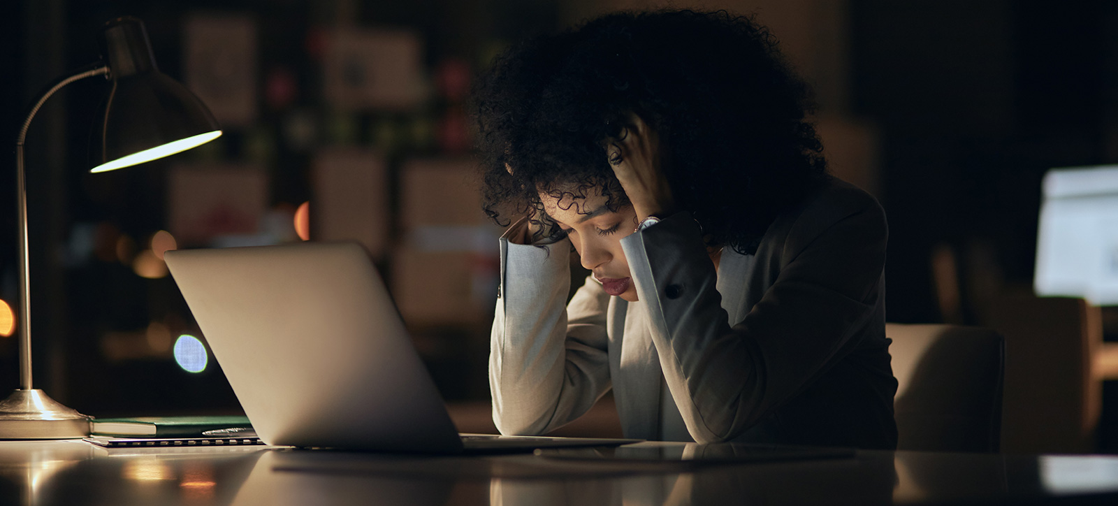 A stressed employee sits at her desk with her head in her hands while working late in a dark office.