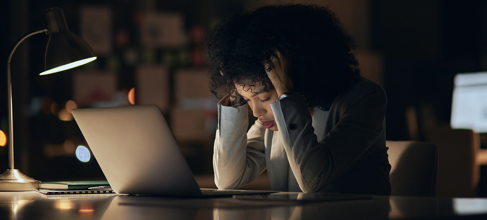 A stressed employee sits at her desk with her head in her hands while working late in a dark office. A stressed employee sits at her desk with her head in her hands while working late in a dark office.