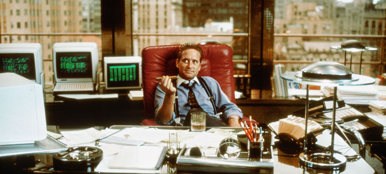 An image of Michael Douglas in his Oscar-winning role as Gordon Gekko in Wall Street. He is sat in a padded, red leather chair and is holding a cigar. A glass of whiskey rests on the desk in front of him. The New York skyline is visible through the window behind him.