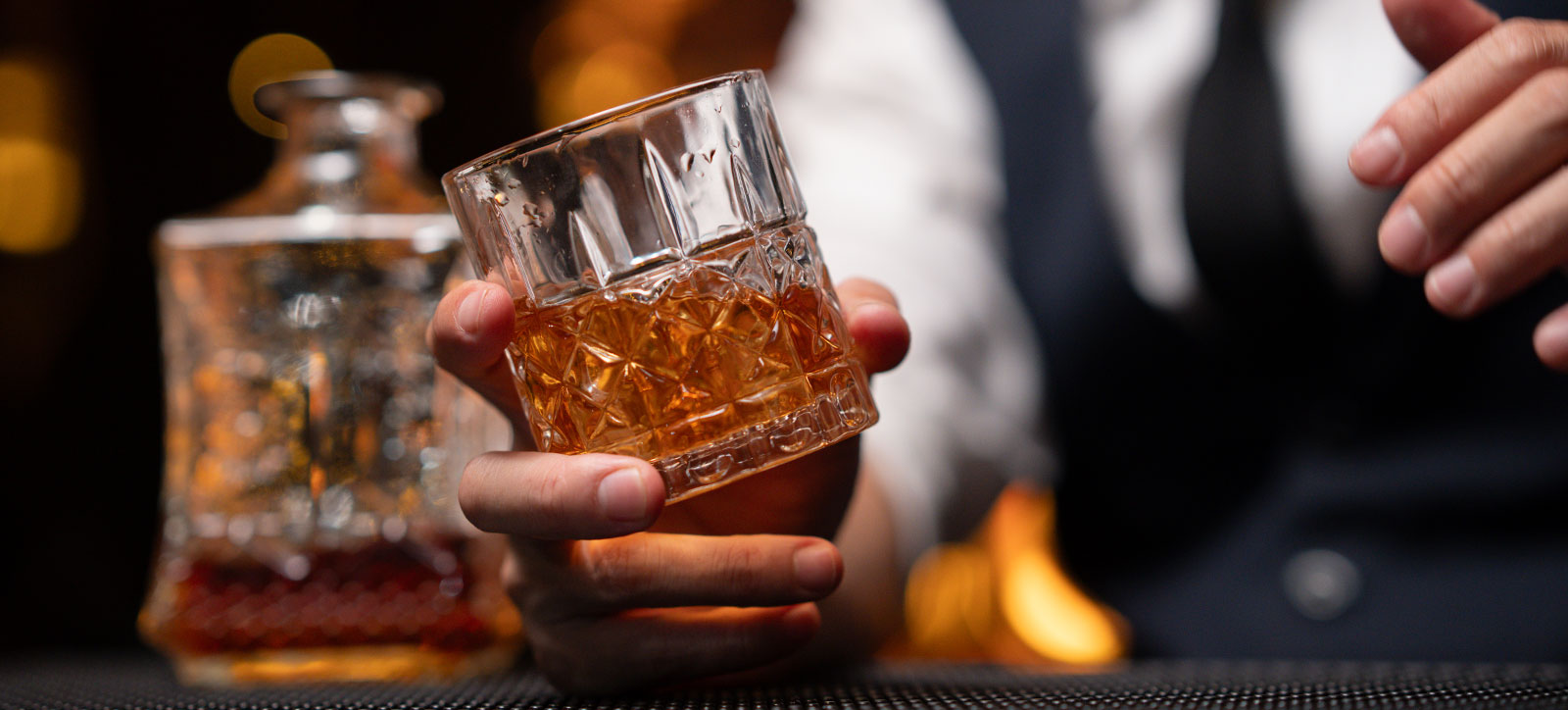 A close up photograph of a bartender's hand holding a glass of whisky. In the background, we can see the decanter the whiskey has been poured from, and the bartender's torso. He is wearing a wear shirt, black tie, and black waistcoat.