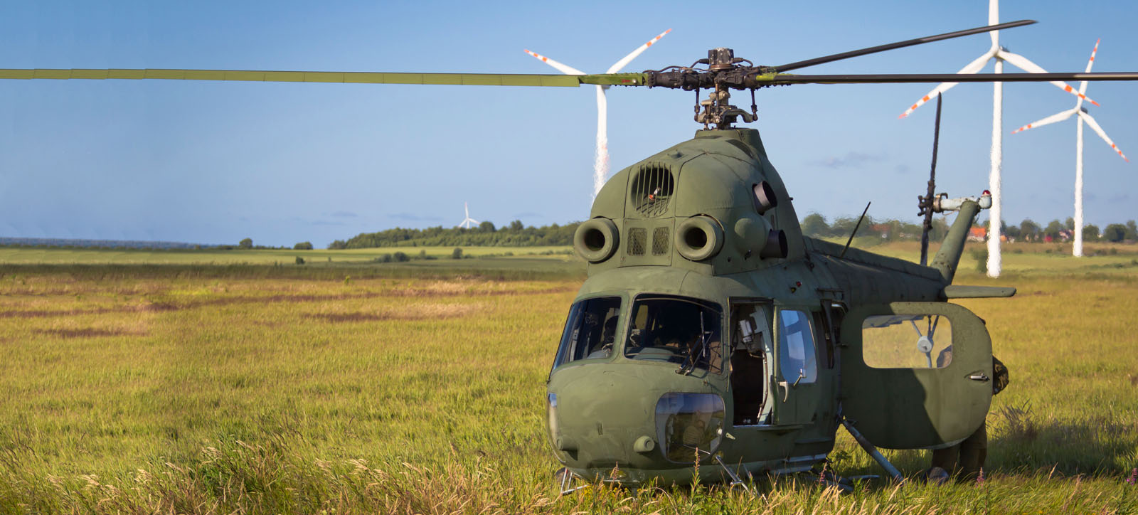 A military helicopter lands in a field in front of a wind farm