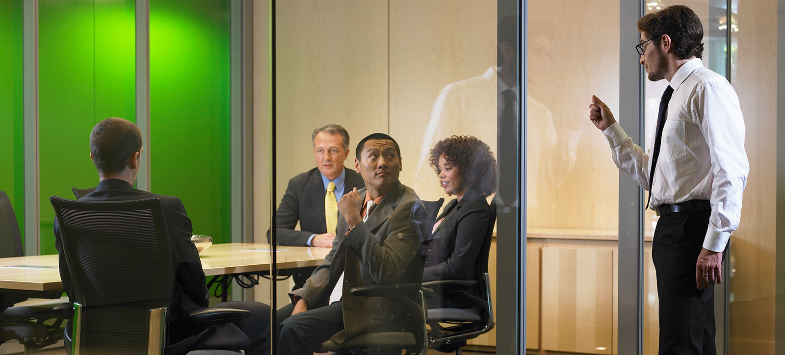 A manager knocks on the door of a glass walled meeting room. Inside, four people are sat around a table, while the rest seem uninterested by his prescence. conducting a meeting without him. One looks over their shoulder at the door