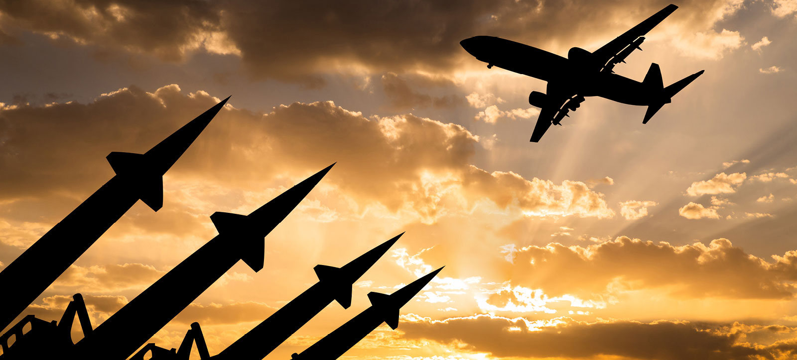 A passenger airplane flies above a battery of military missiles pointed towards the sky. Both the plane and the missiles are silhouetted against an orange sunset and dark clouds.
