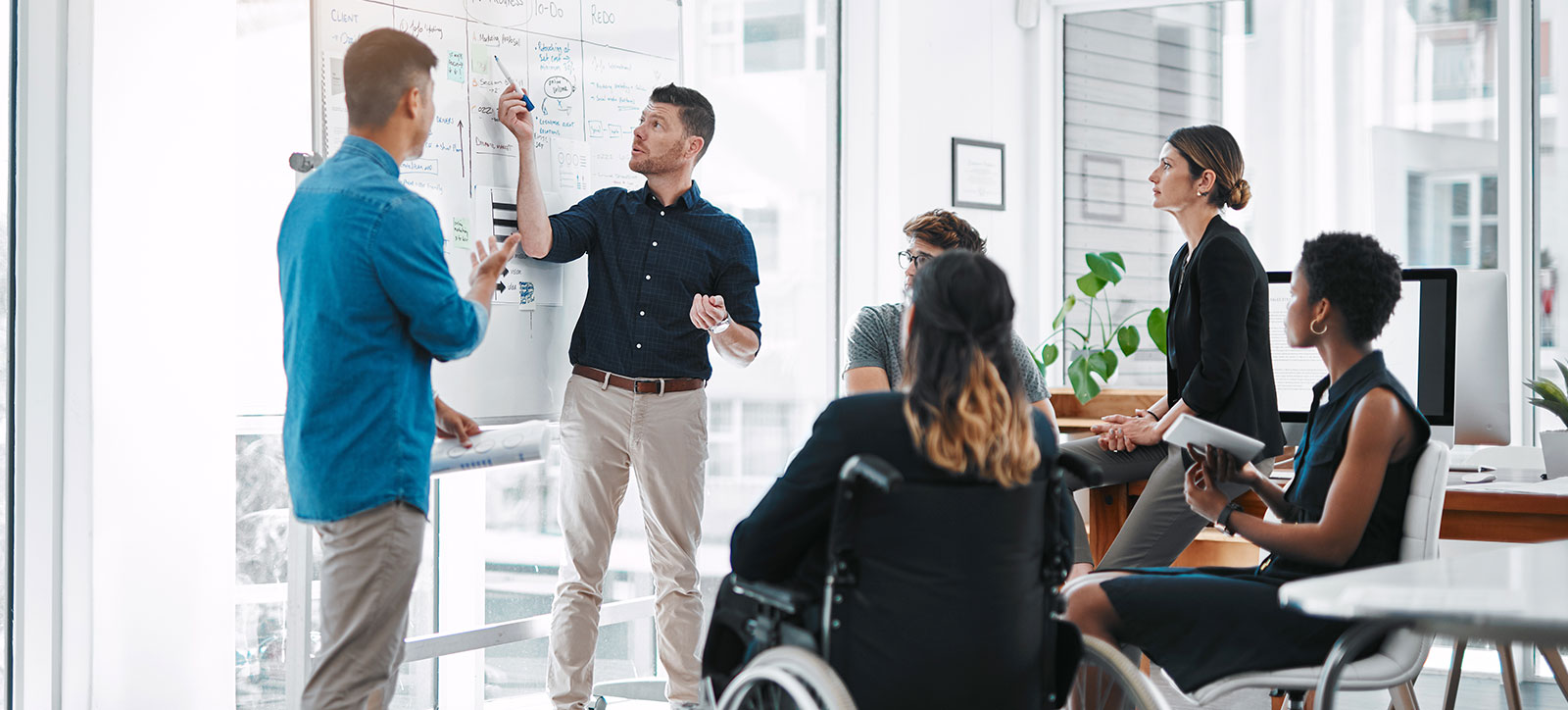 six staff including one in a wheelchair around a whiteboard as a man explains the idea