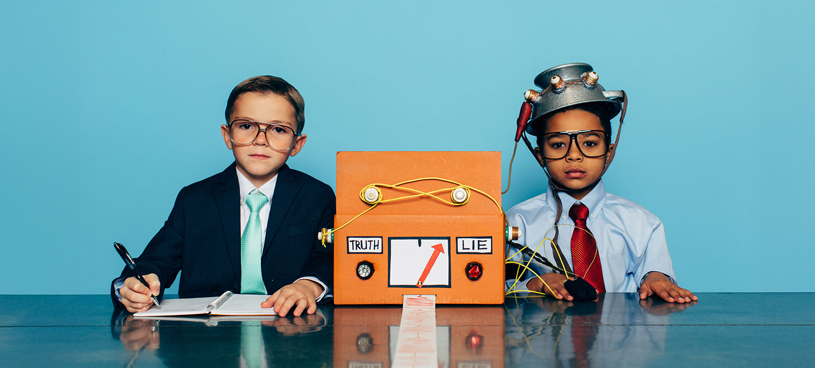 Two boys sat side by side at a desk. Both are dressed as office workers, wearing shirts and ties. One is fitted to a homemade, pretend lie detector. He has an upturned colander on his head, with wires connected it to a decorated cardboard box. A long strip of paper with a red polygraph line drawn on is emerging from the box. The other boy is wearing a suit jacket to make him look more like an authority figure and has a pen in his hand and is recording the results in a notebook.
