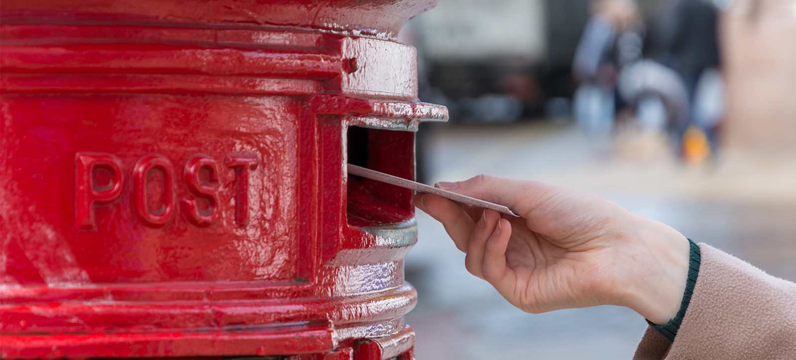 A close up of a woman's hand post a small stack of letters into a bright red Royal Mail postbox. The woman is wearing a dark, woolen sweater and a beige coat. The high street in the background is in soft focus, making it impossible to see individual details.