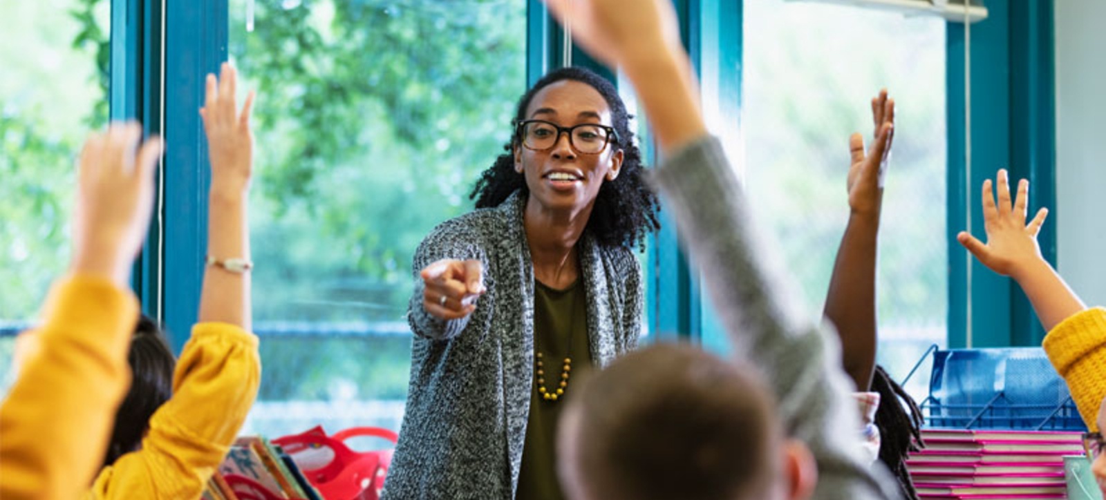 A female primary school teacher chooses who will answer a question from a group of children with their hands raised.
