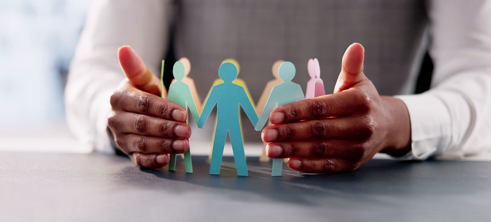 A close up of a leader's hands protectively surrounding a circle of different coloured paper figures of people, representing how organisations can foster inclusive leadership. A close up of a leader's hands protectively surrounding a circle of different coloured paper figures of people, representing how organisations can foster inclusive leadership.