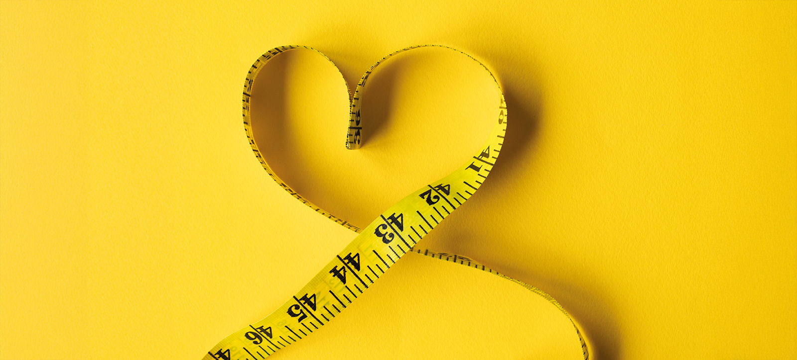 A traditional yellow, metal tape measure is looped around to form a heart shape. It is lying on a bright yellow background. A traditional yellow, metal tape measure is looped around to form a heart shape. It is lying on a bright yellow background.