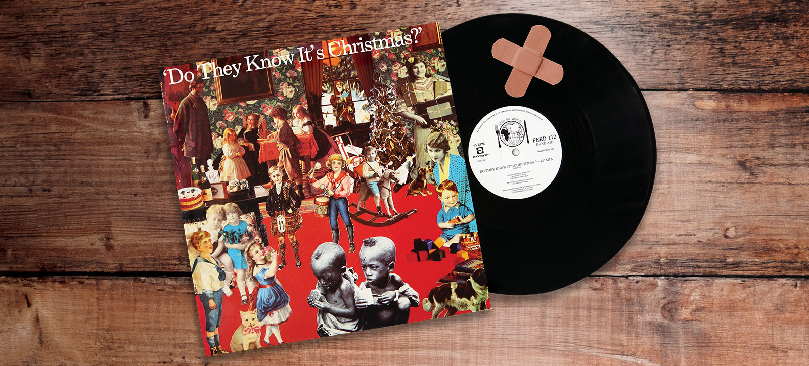A vinyl LP of Band Aid's charity single Do They Know It's Christmas? slips out of its carboard sleeve onto a tabletop. Two plasters are stuck to the LP forming an x-shaped cross
