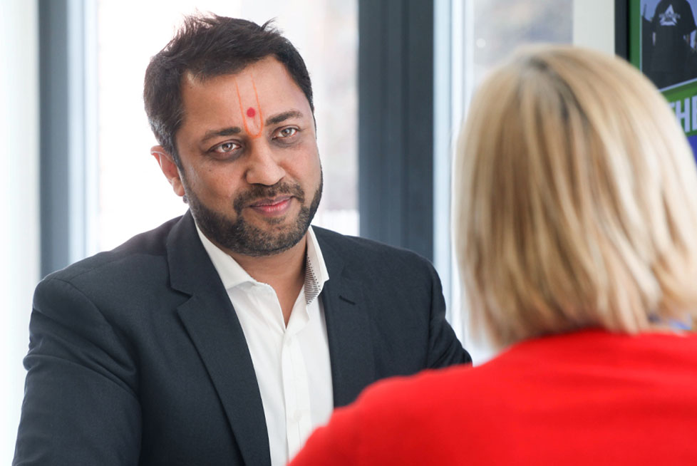A man with a tilak on his forehead and a suit speaks to a woman with blonde hair wearing a red top in an office setting