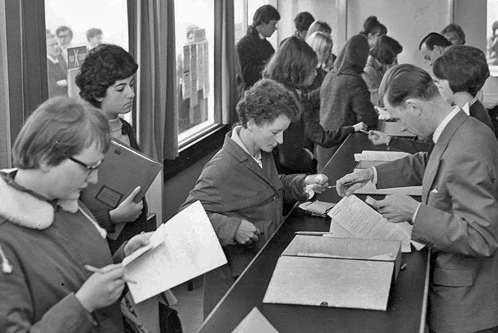 A group of individuals waiting at a reception desk, reviewing documents and engaging in discussion.