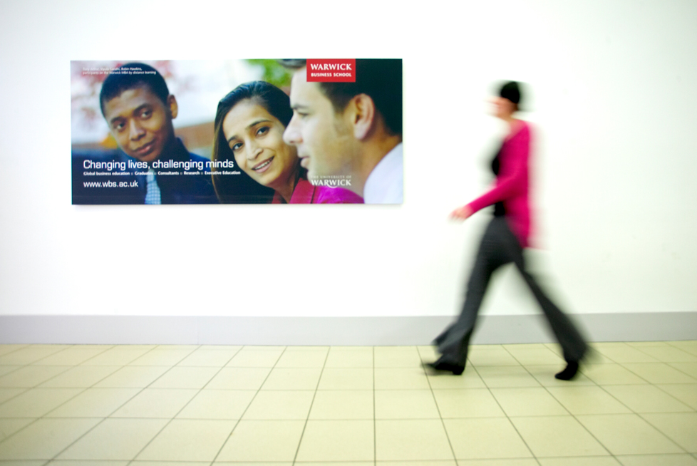 A women walking past a billboard that is advertising Warwick Business School